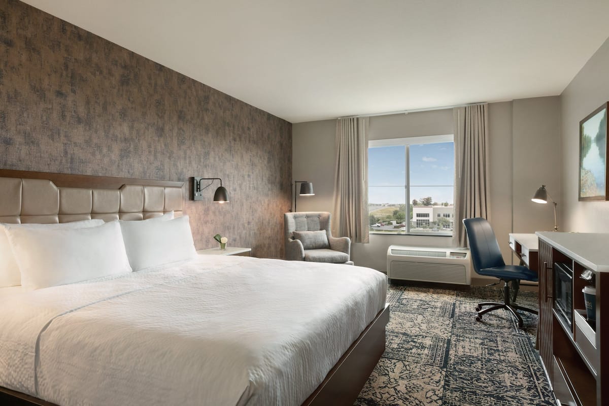 A hotel room with a large bed, a desk, a chair, a TV, and a window with a view of buildings.
