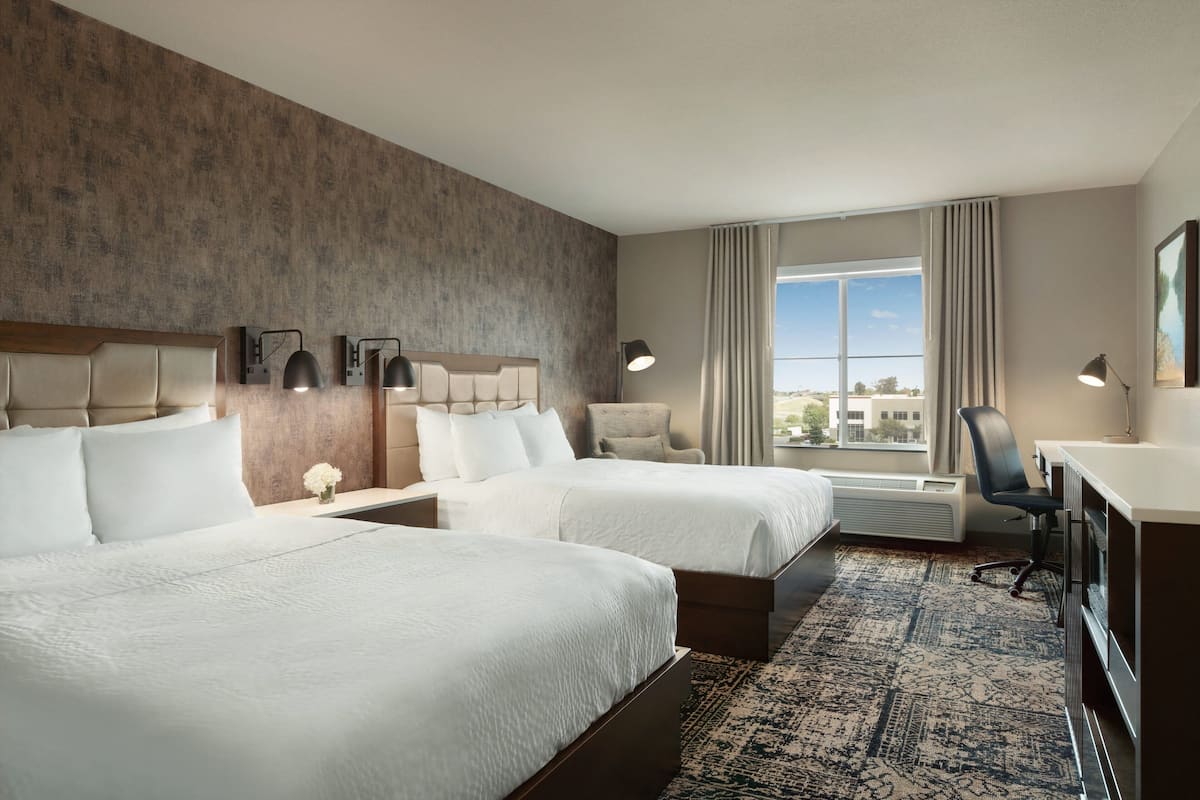 A hotel room with two beds, a desk, a chair, a lamp, and a large window with a view.