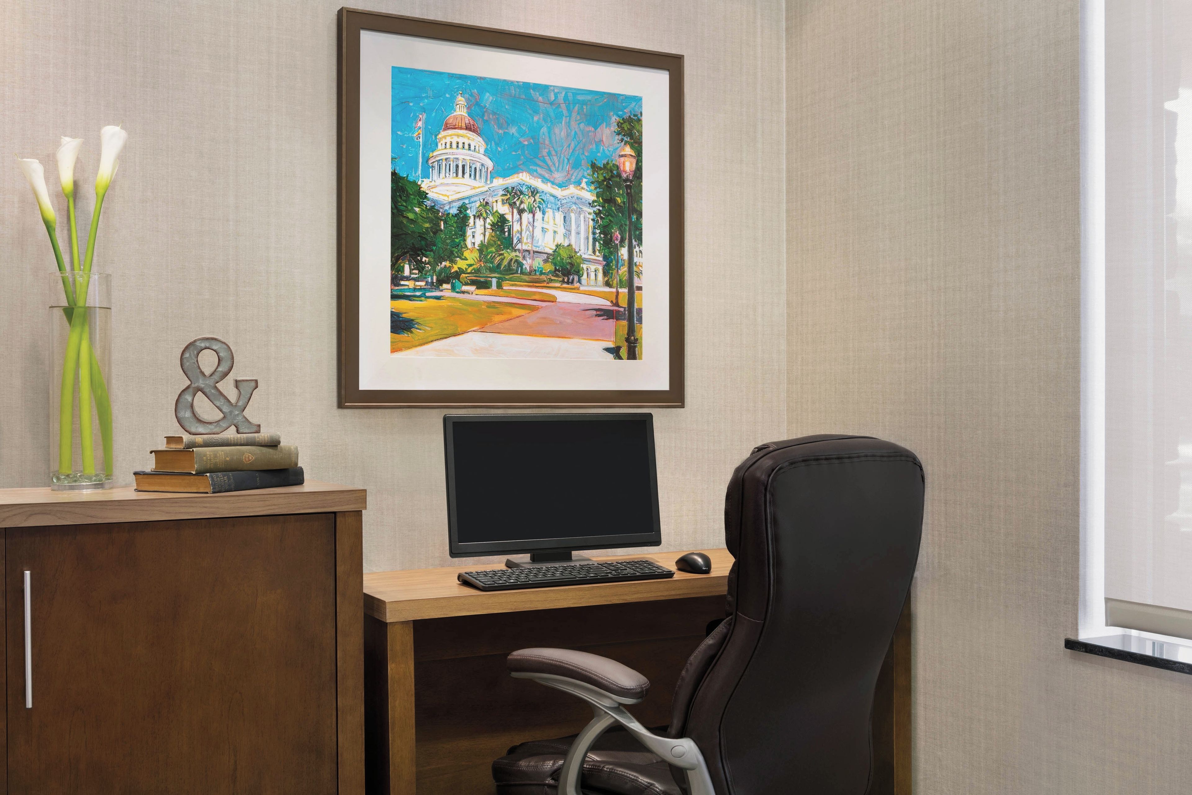 A hotel room with a desk, a computer, and a chair. There is a framed painting of a building on the wall.