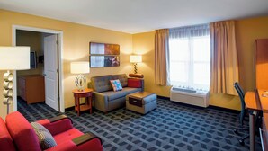 Habitación de hotel con sofá, sillón y una pequeña mesa con lámpara. Se ve una ventana con cortinas y una puerta que da a otra habitación.