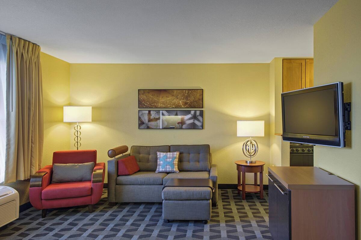 A hotel room with a sofa, armchair, ottoman, flat-screen TV, and a small table with a lamp.