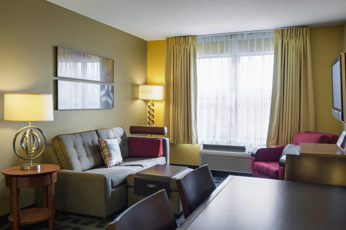 A hotel room with a sofa, a small table, a desk, a TV, and a window with curtains.