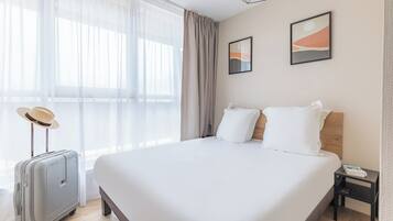 A hotel room with a large bed, a suitcase, a nightstand, and a window with sheer curtains.