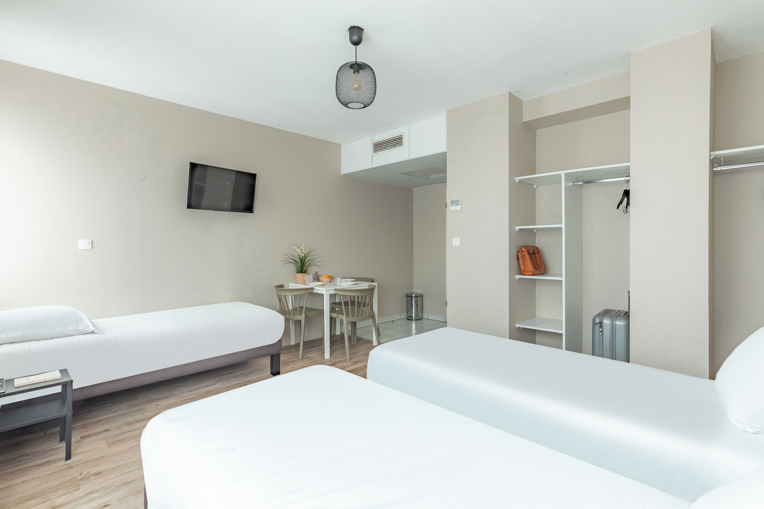 Ein Hotelzimmer mit zwei Einzelbetten, einem Schreibtisch mit Stuhl, einem kleinen Tisch mit Pflanze, einem an der Wand montierten Fernseher und einem Kleiderschrank mit Kleiderstange.
