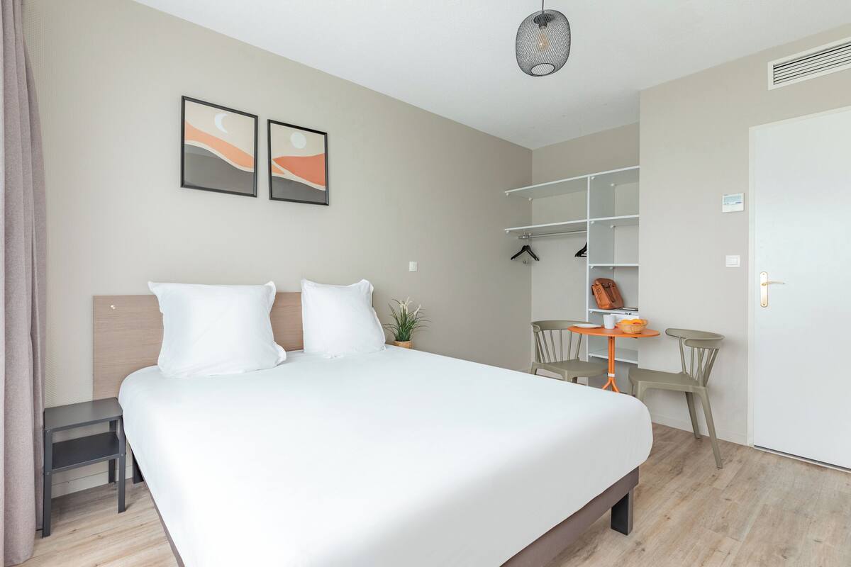 Una habitación de hotel moderna con una cama grande, un escritorio pequeño y una silla. Hay una planta en el escritorio y un estante con ropa.
