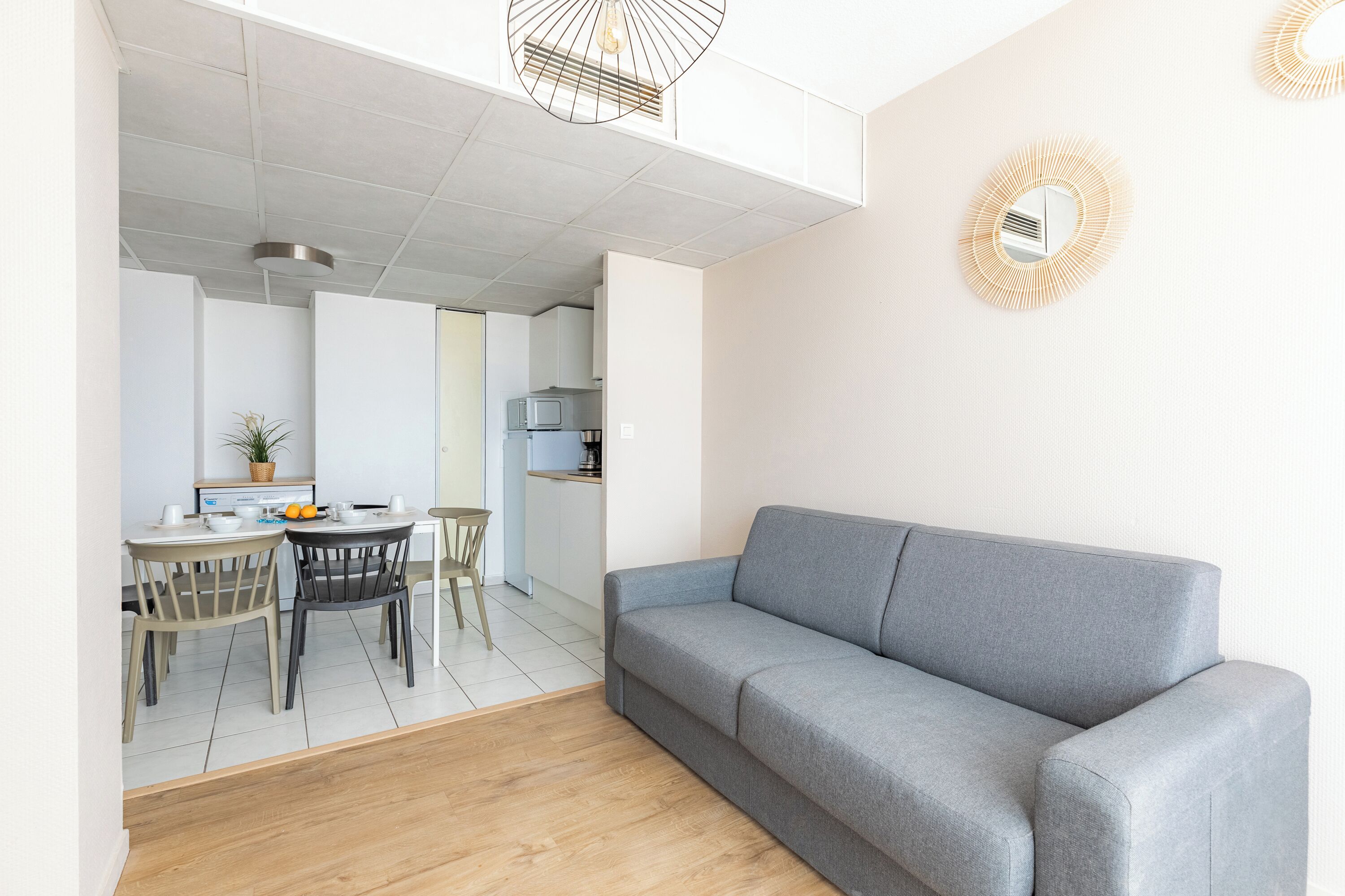 Un salon moderne avec un canapé gris, une table à manger en bois et des chaises. On aperçoit une cuisine à l’arrière-plan.