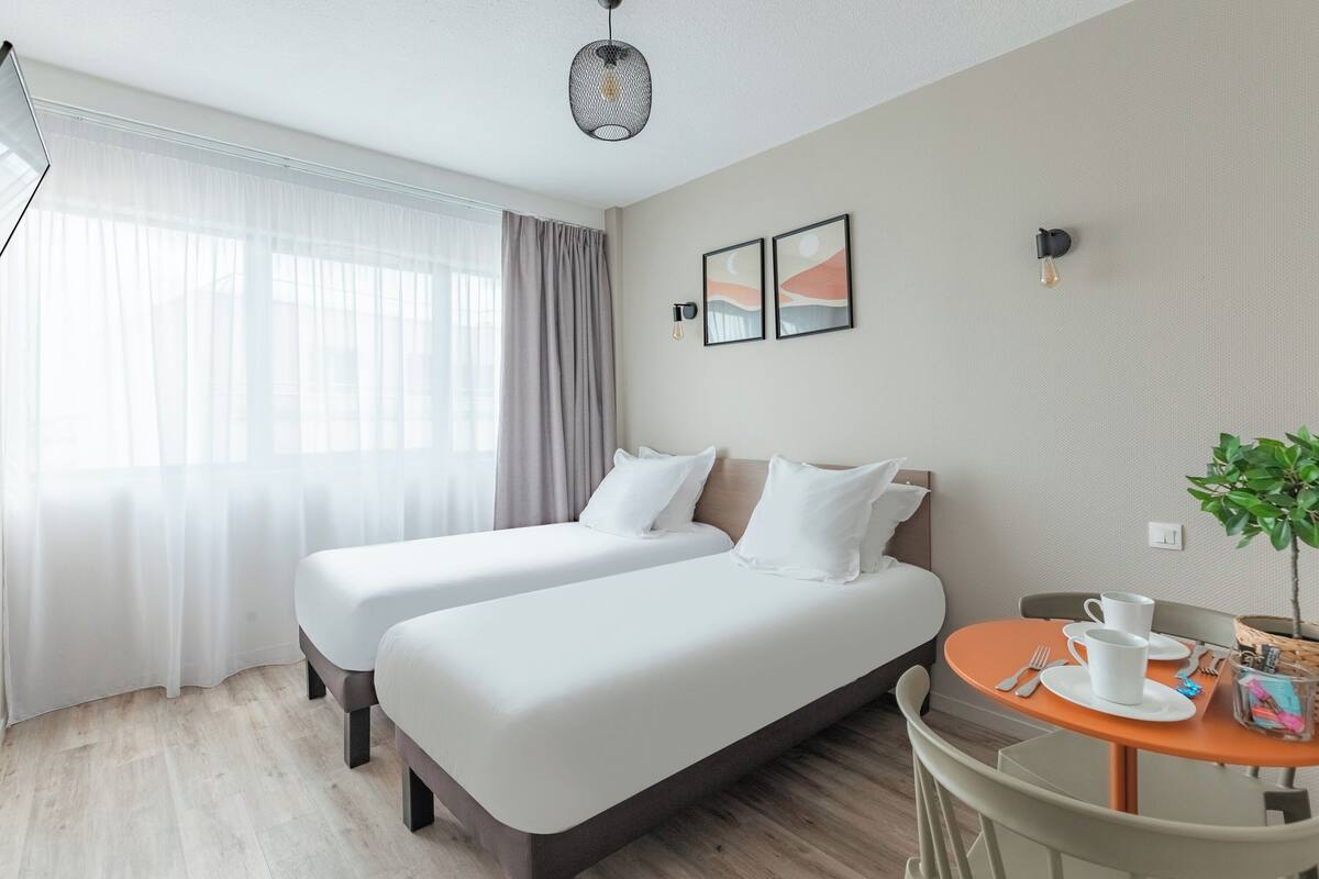 Habitación de hotel con dos camas, una mesa de comedor con sillas, una mesita con una planta y obras de arte en la pared.