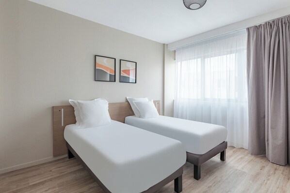A hotel room with two single beds, a wooden headboard, a large window with sheer curtains, and two framed abstract paintings on the wall. - Appart'City Classic Bordeaux Centre (Bordeaux)