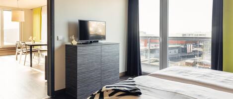 Ein modernes Hotelzimmer mit einem großen Bett, einem Flachbildfernseher auf einer dunklen Holzkommode und Blick auf die Stadt durch große Fenster.