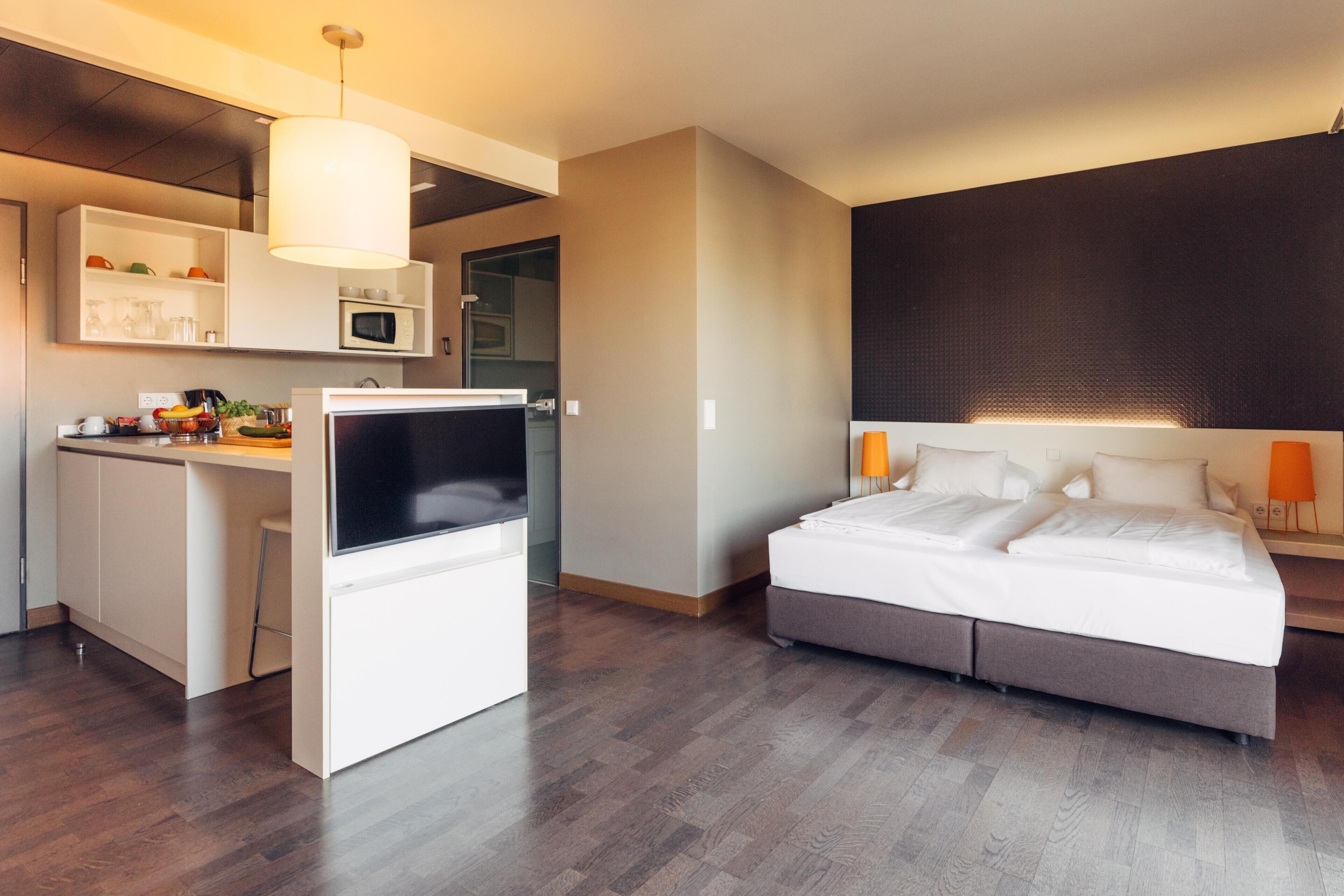 Kompakti hotellihuone, jossa on pieni keittiönurkkaus, sänky ja suuri ikkuna.