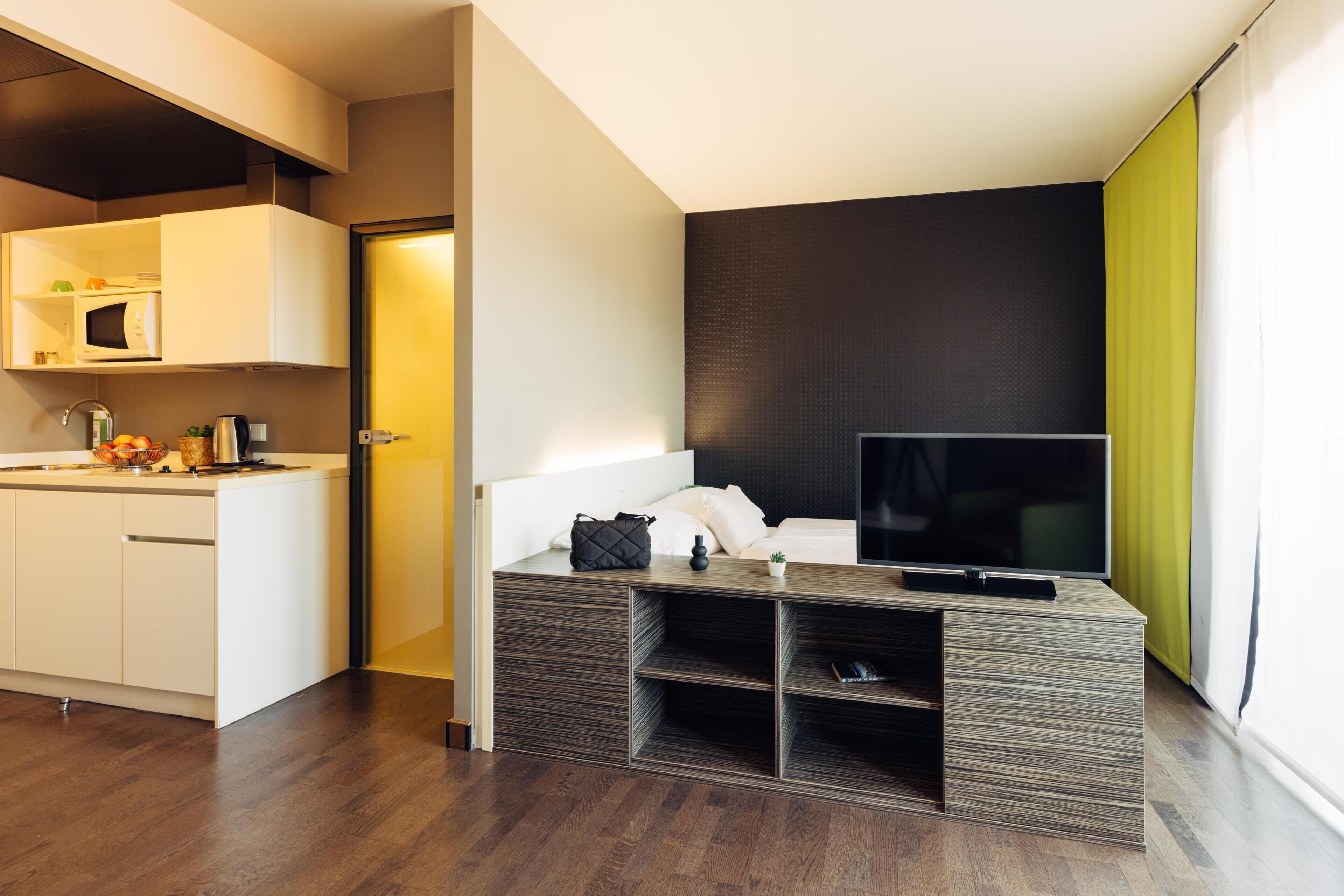 Ein modernes Hotelzimmer mit einer Küchenzeile, einem Flachbildfernseher, einem Bett und einem großen Fenster.