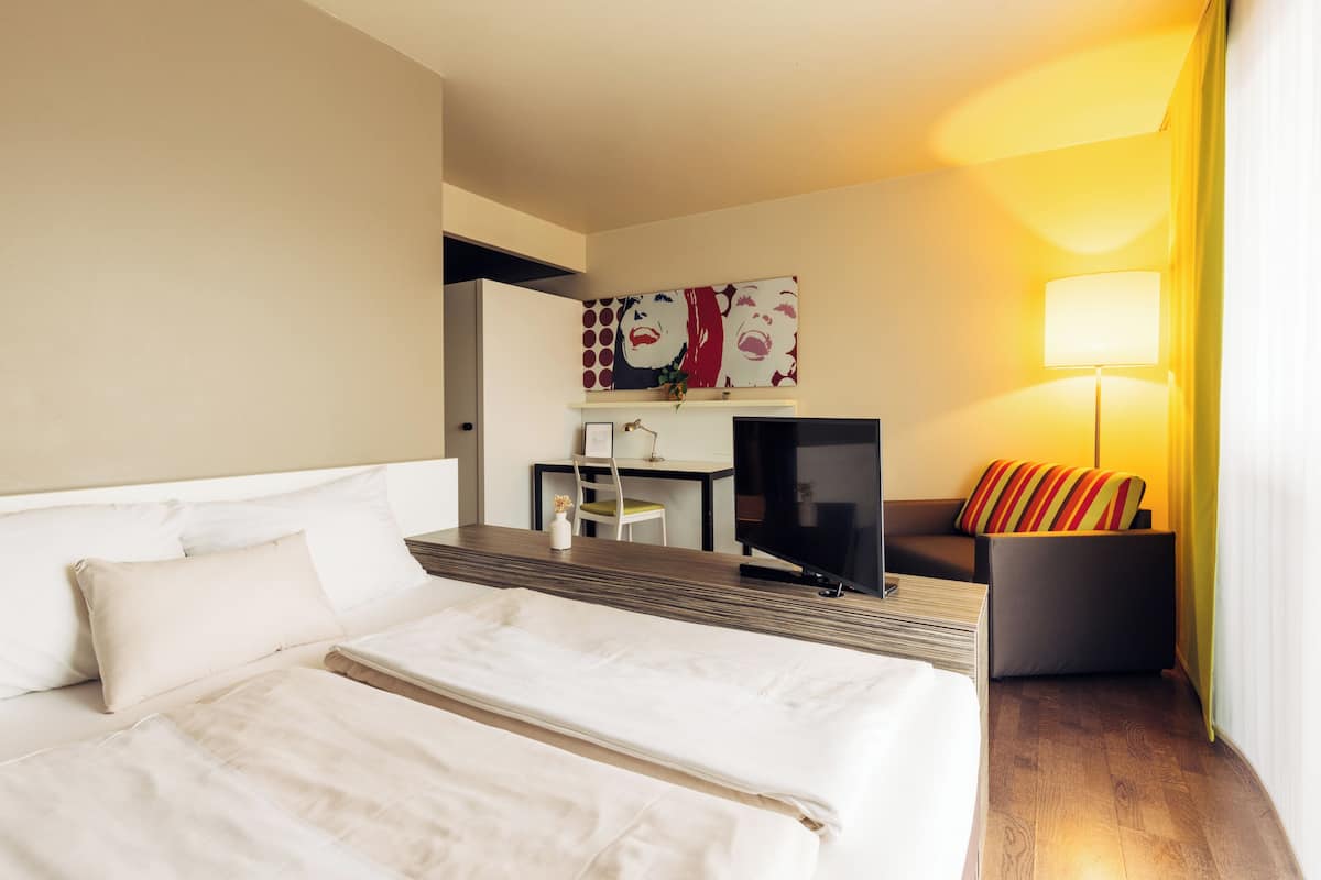 A hotel room with a large bed, a desk with a chair, a television, and a sofa with a colorful cushion.