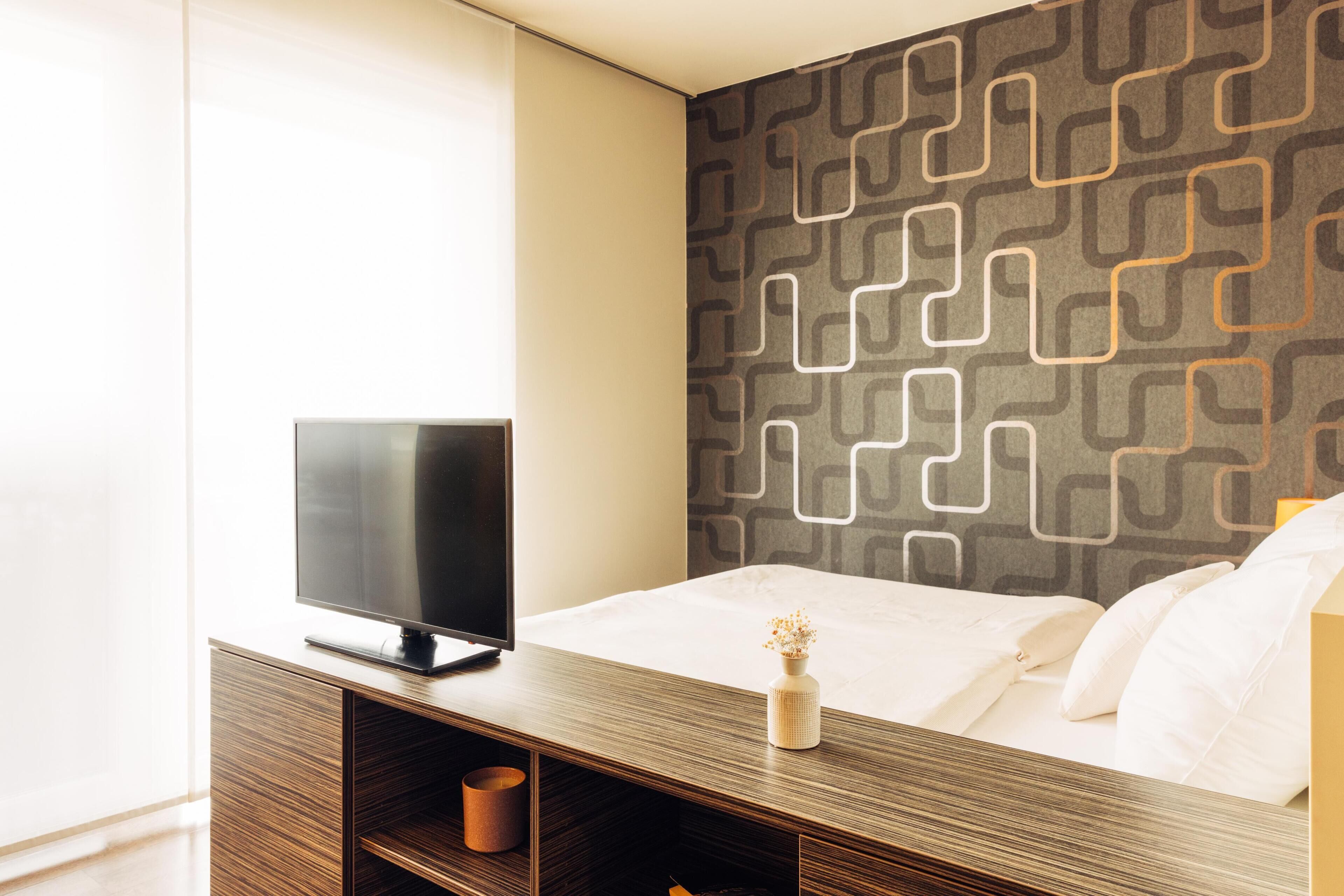 A modern hotel room with a bed, a TV on a wooden console, and a patterned wall.
