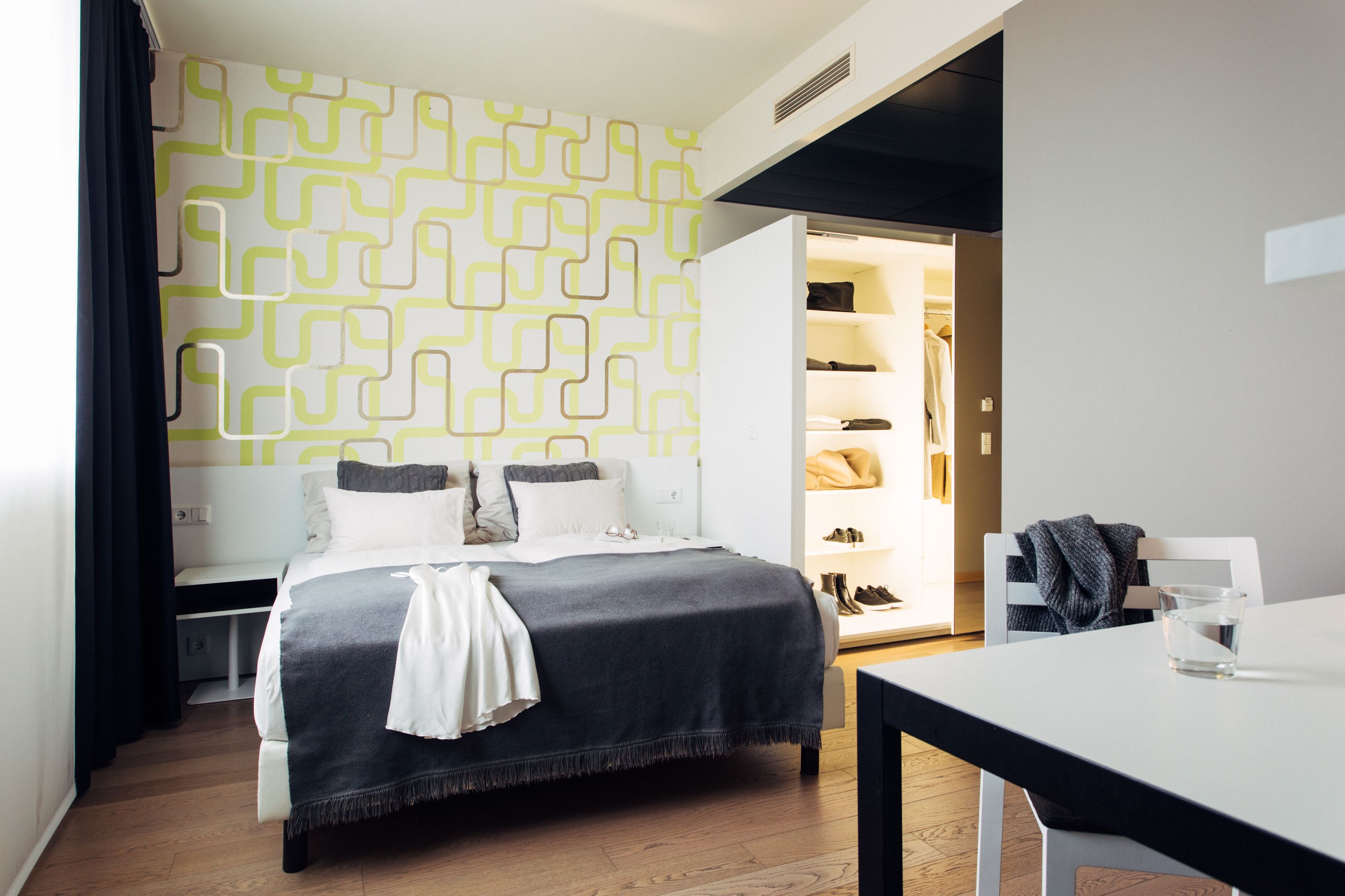 A modern hotel room with a neatly made bed, a small table, and a wardrobe with hanging clothes.