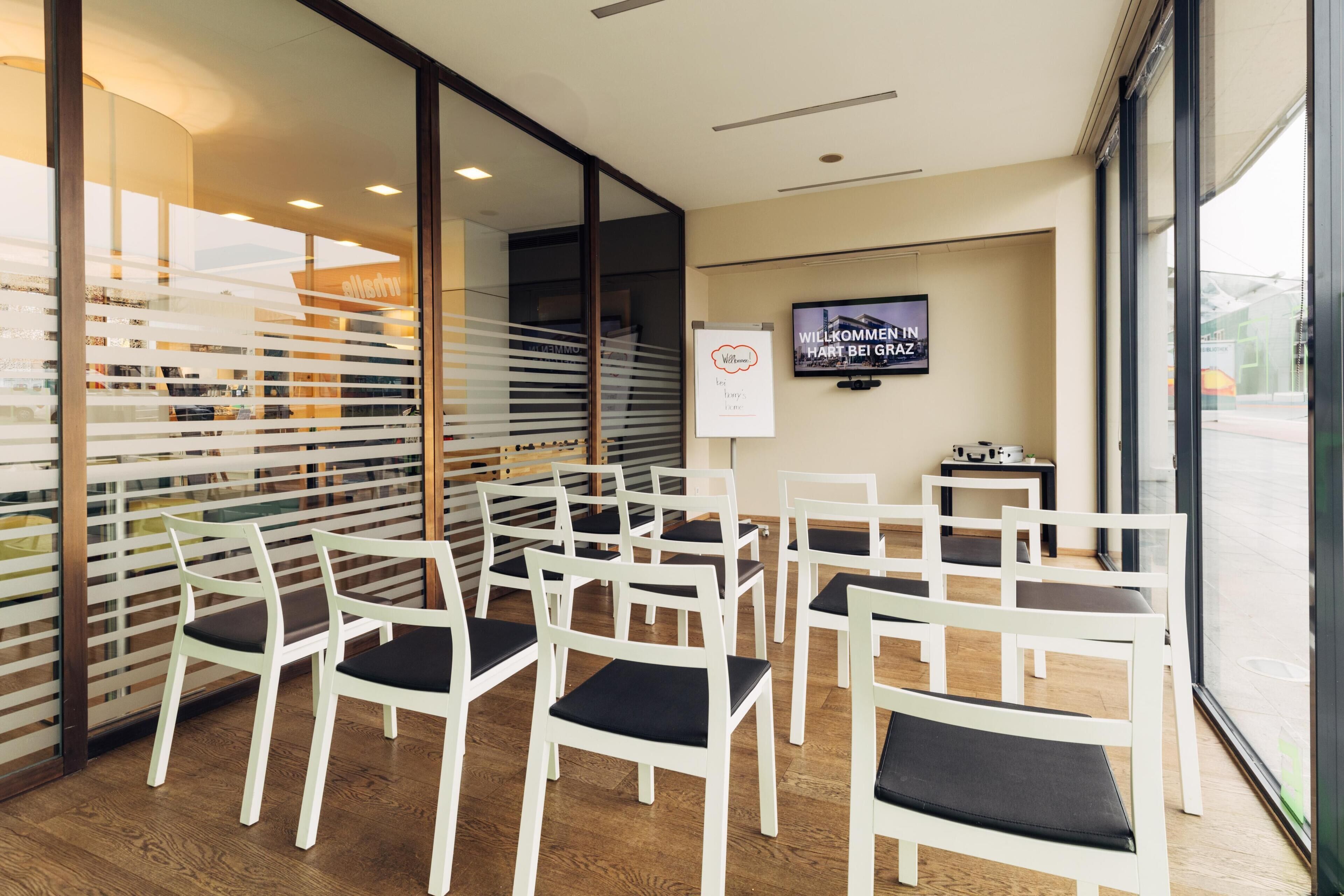 A room with white chairs and tables, a TV on the wall, and a sign that reads "WILLKOMMEN IN HART BEI GRZ".