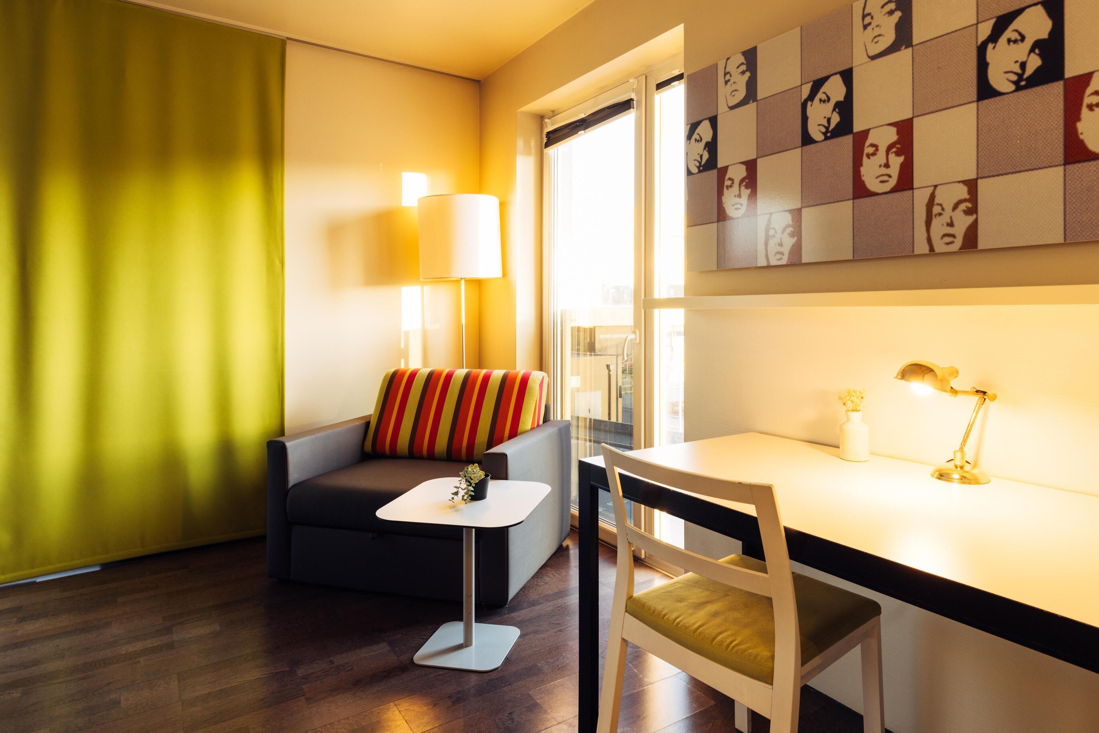 A modern living room with a sofa, a small table, a desk with a chair, a lamp, and a wall with artwork.