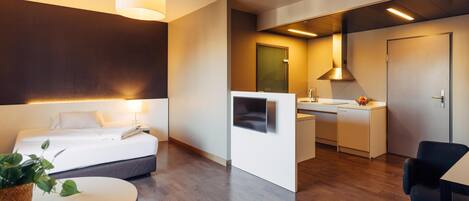 A modern hotel room with a bed, bedside table, and a small kitchen area.