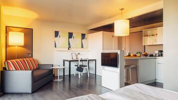 Quarto de hotel moderno com sofá, mesa pequena, televisão, cozinha compacta e área de refeições.