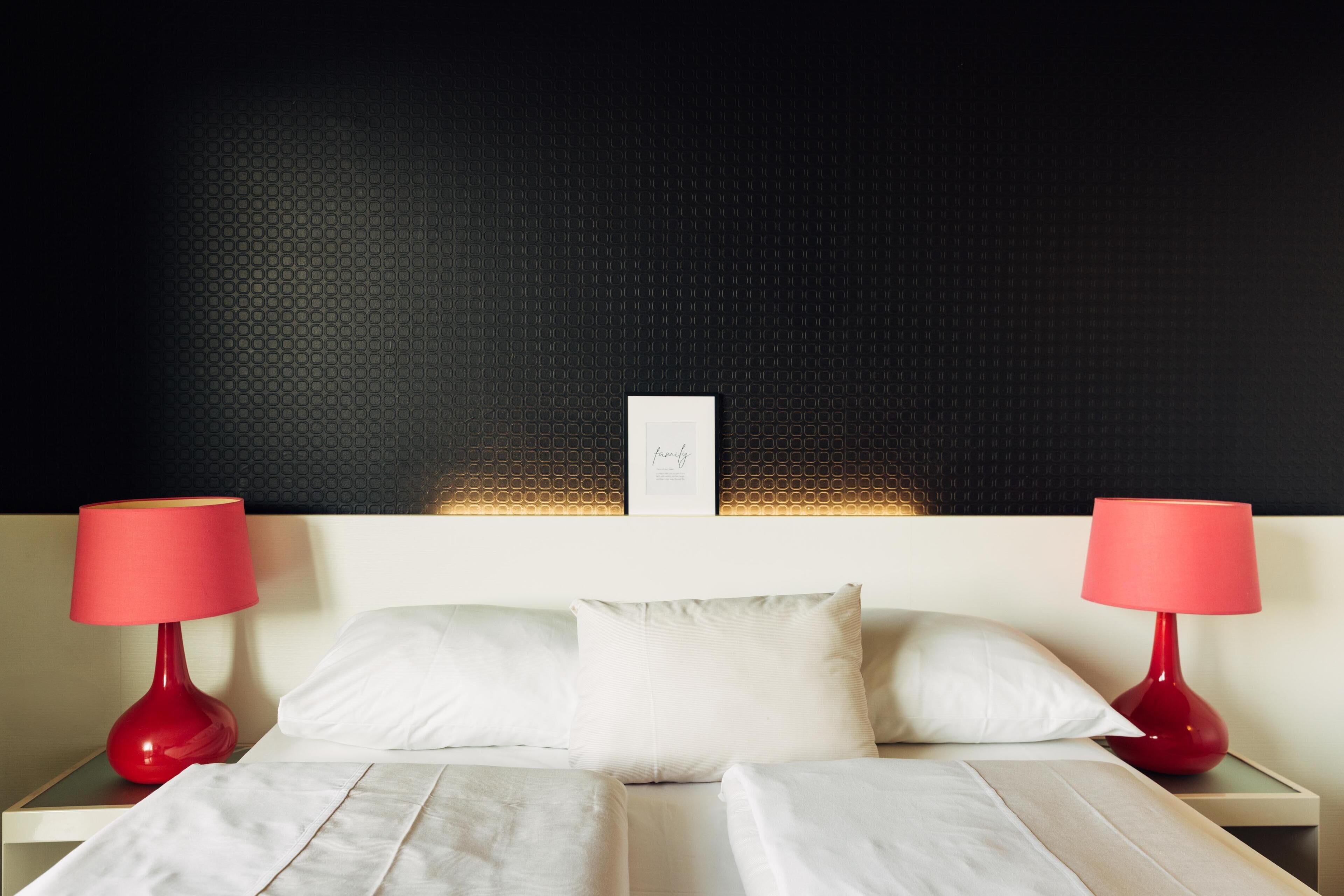 Een net opgemaakt bed met witte lakens, twee rode bedlampen en een zwart gestructureerde muur op de achtergrond.