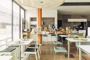 A modern cafe with white tables, green chairs, and a central counter with various food items and a chalkboard menu. - harry’s home Hart bei Graz hotel & apartments (Hart bei Graz)