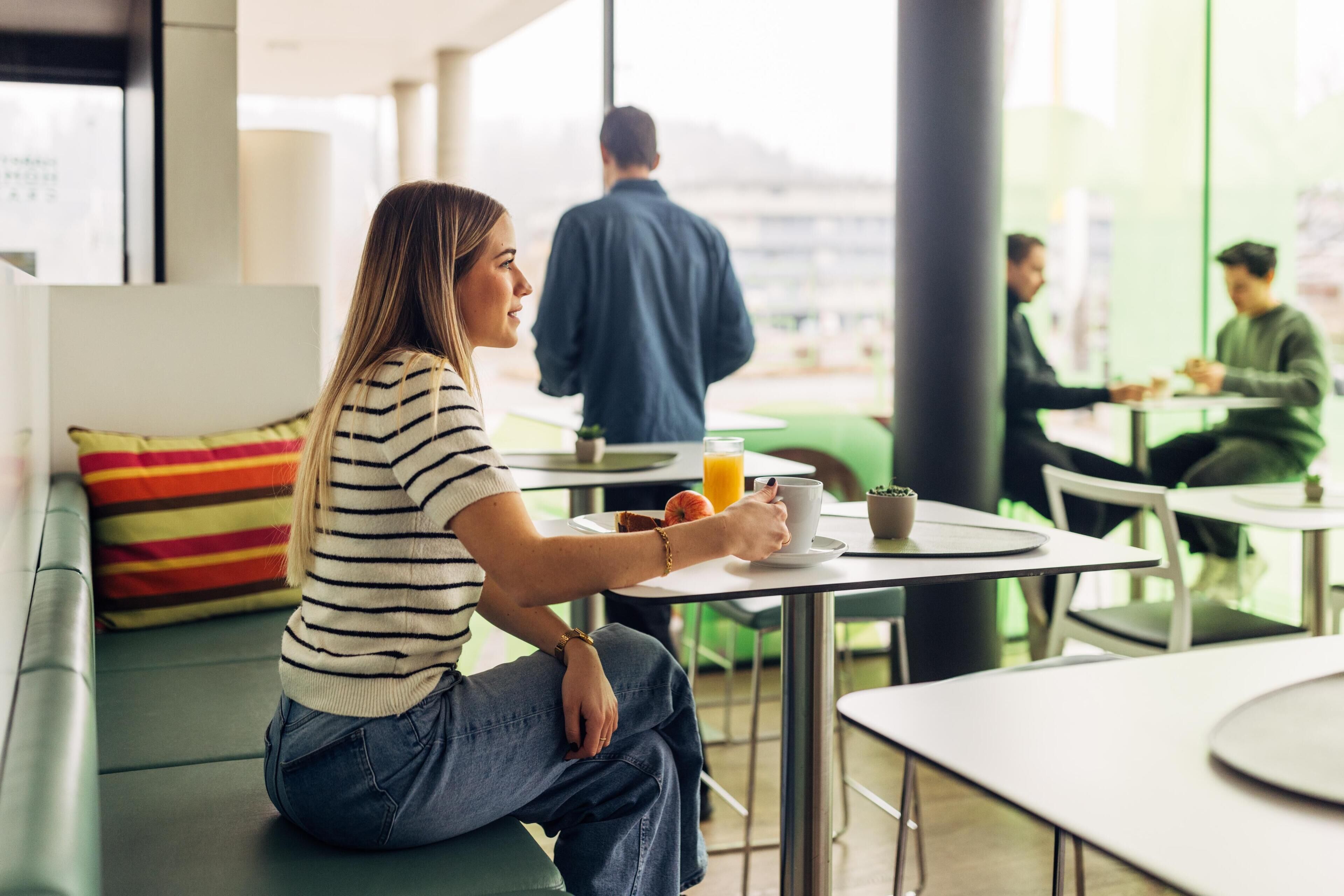 Une personne assise sur un canapé vert, avec un coussin à rayures, tenant une tasse et une soucoupe.