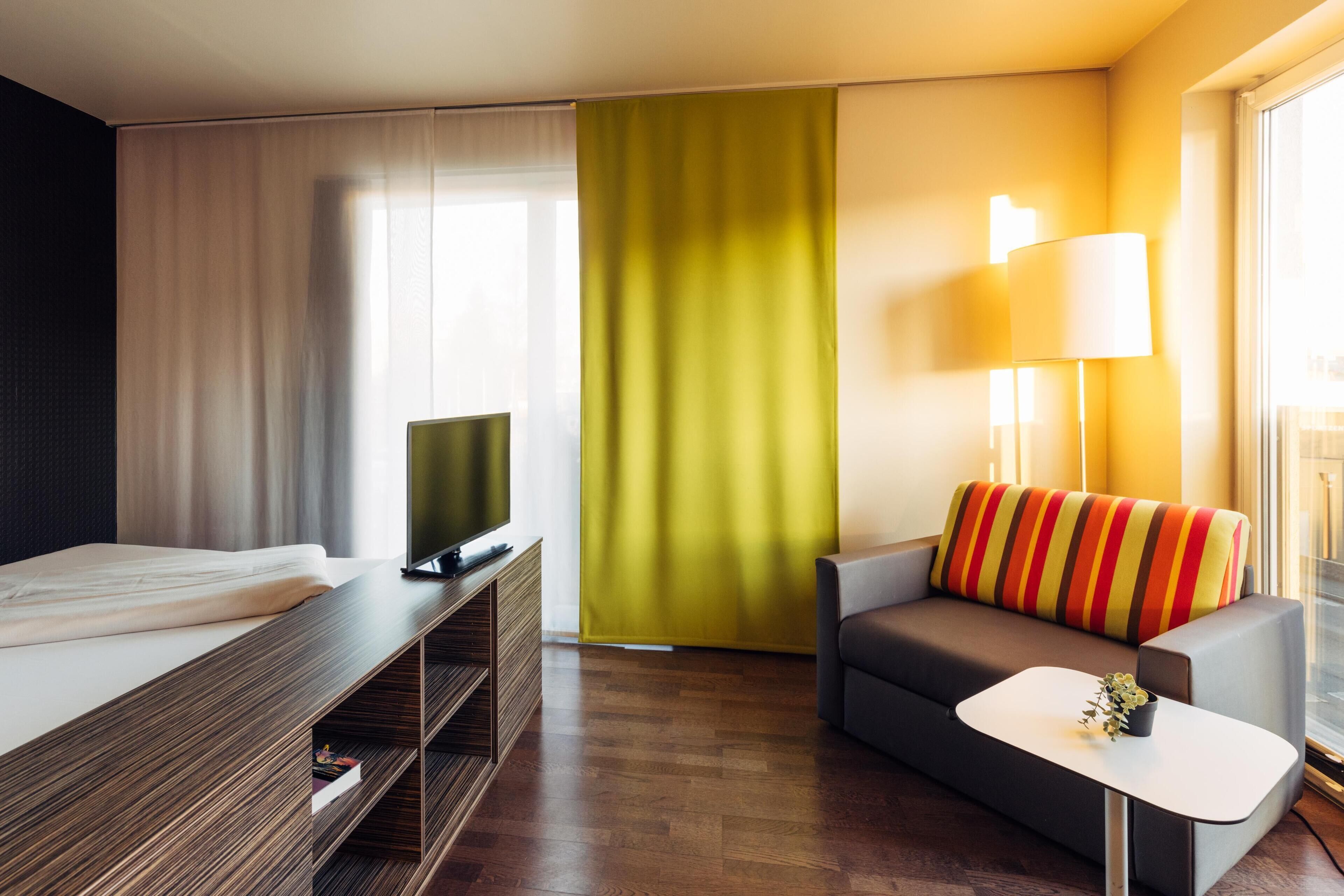 Una habitación de hotel moderna con una cama, un televisor sobre un soporte de madera, un sofá con un cojín de colores, una mesita de noche con una lámpara y un ventanal con cortinas.