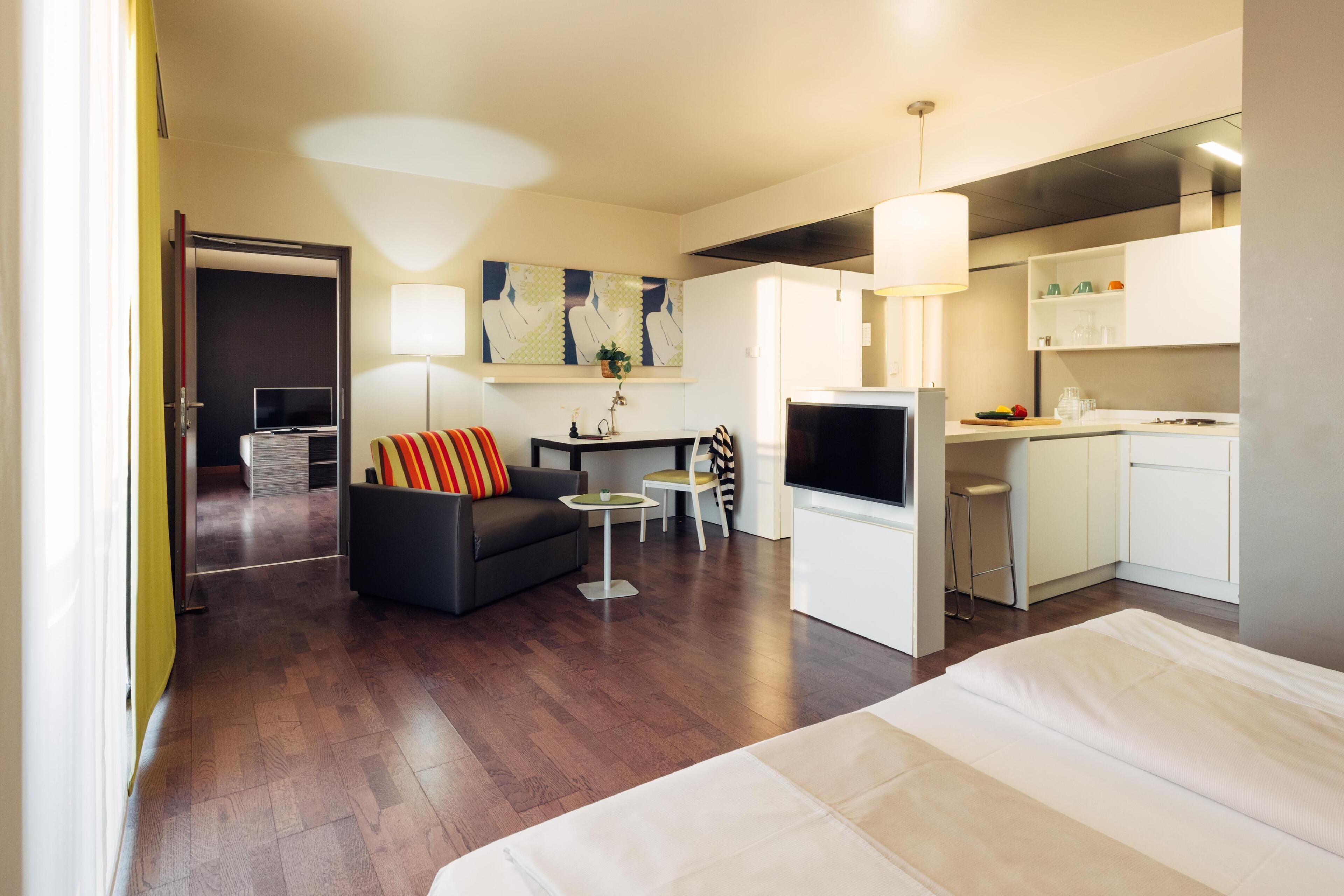 A modern hotel room with a bed, a sofa, a TV, a small table, and a kitchen area.