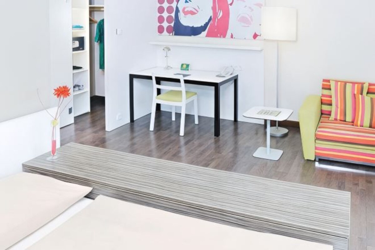 A modern room with a dining table, a chair, a small white table, a colorful sofa, and a painting on the wall.