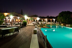 An outdoor pool area with tables and chairs, surrounded by a wooden deck and a covered seating area with lights. - Olympic Village Hotel & Spa (Archaia Olympia)