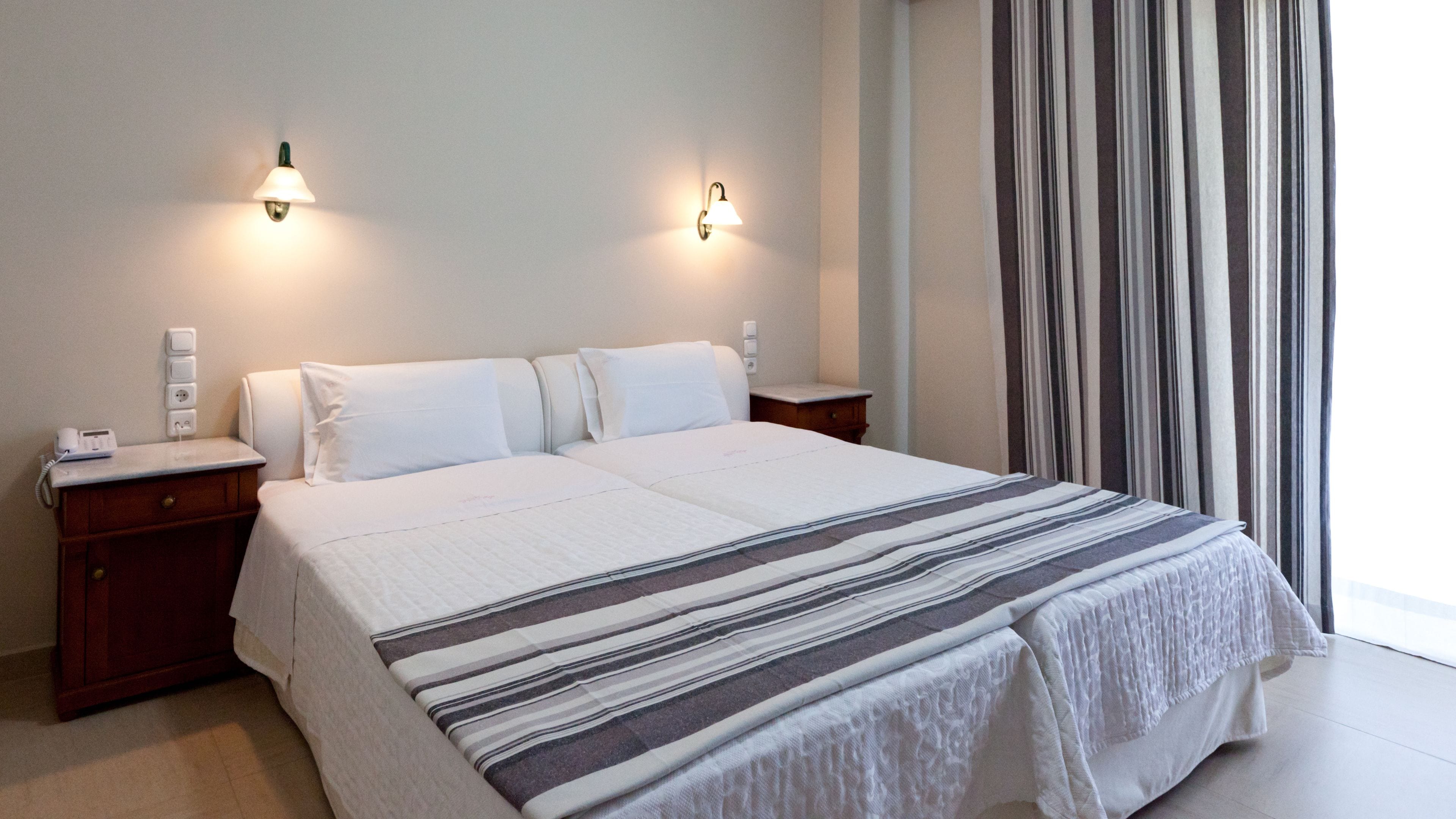 A hotel room with a large bed, two bedside tables, a telephone, and a window with striped curtains.