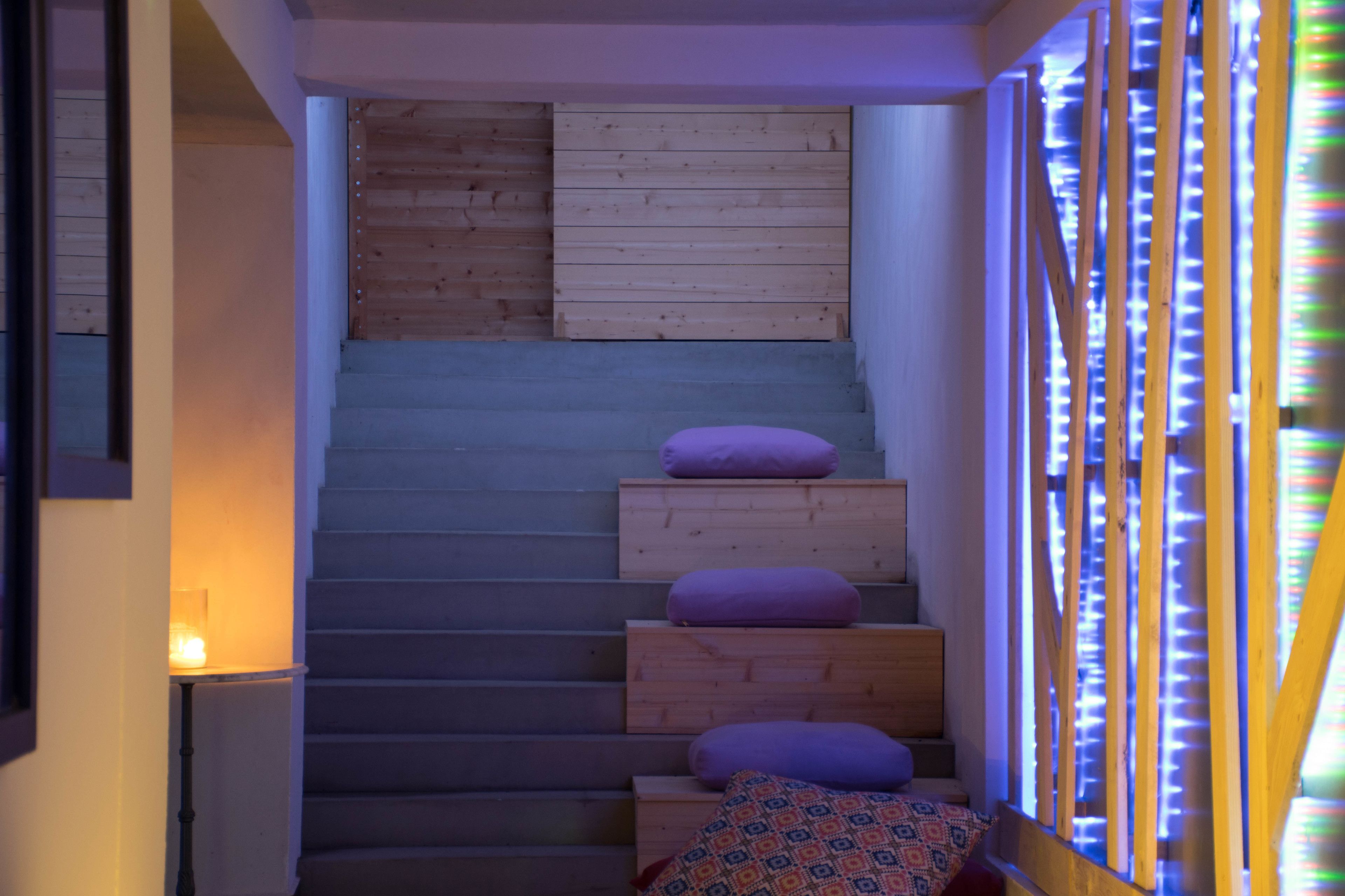 Een trap met houten treden en kussens, leidend naar een kamer met een houten wand en een raam met verticale lamellen.