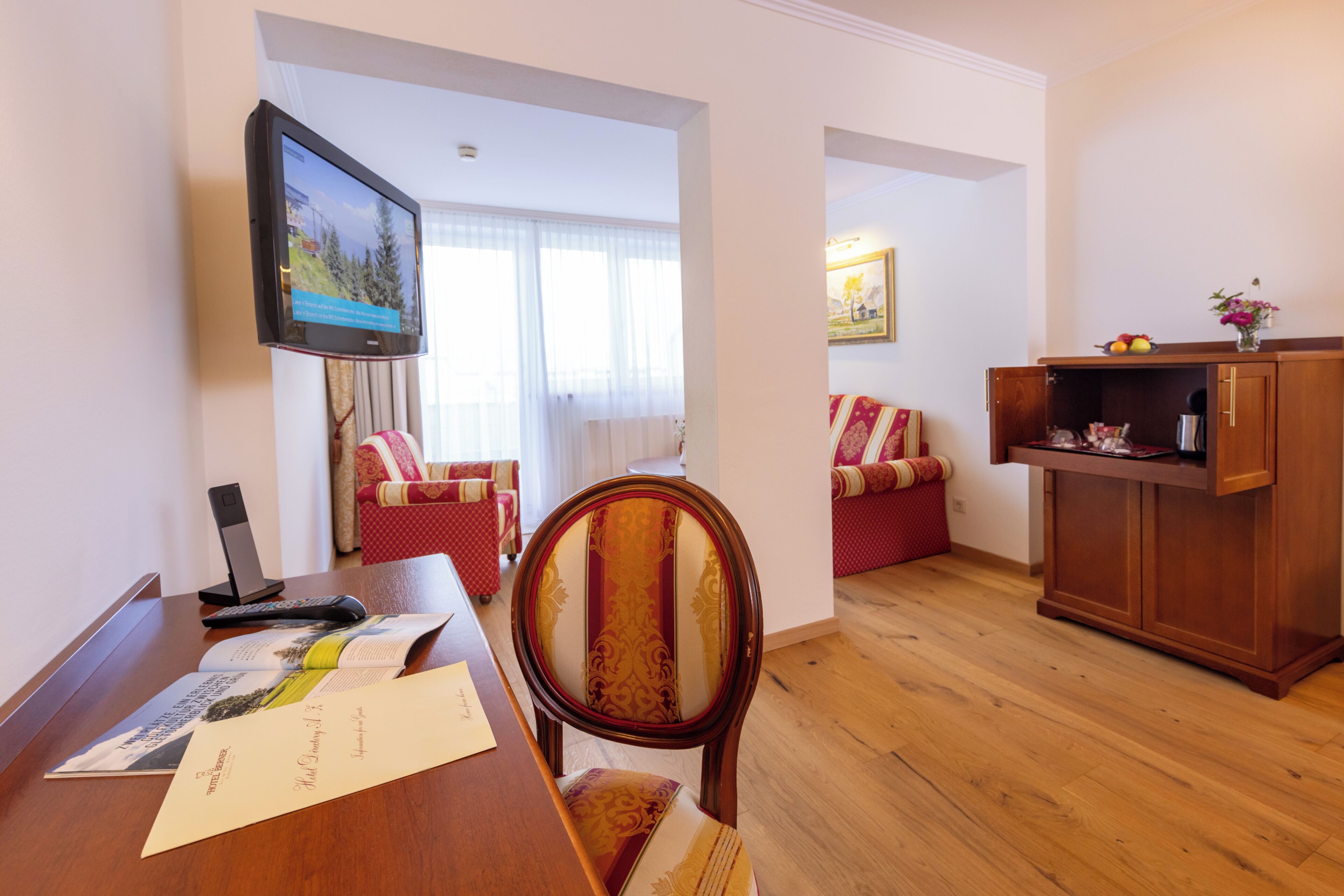 A hotel room with a wooden desk, a television mounted on the wall, a red and white patterned sofa, and a wooden cabinet with a vase of flowers.