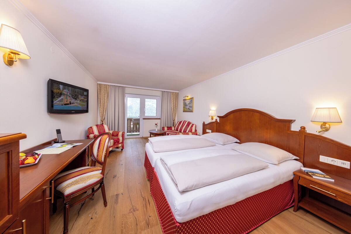 Une chambre d’hôtel avec un lit en bois, un bureau avec une chaise, une télévision et une fenêtre avec des rideaux.