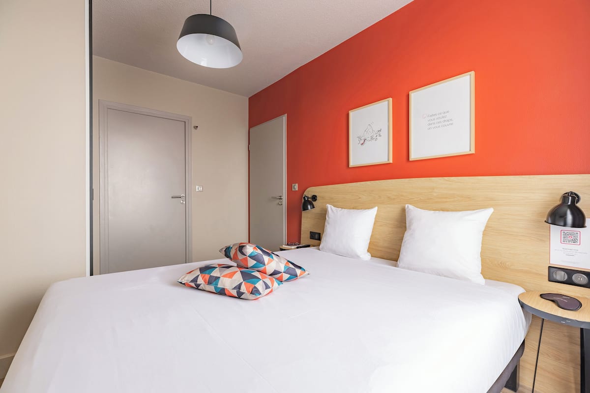 Quarto de hotel com cama, almofadas, mesa de cabeceira com abajur e duas imagens emolduradas na parede.