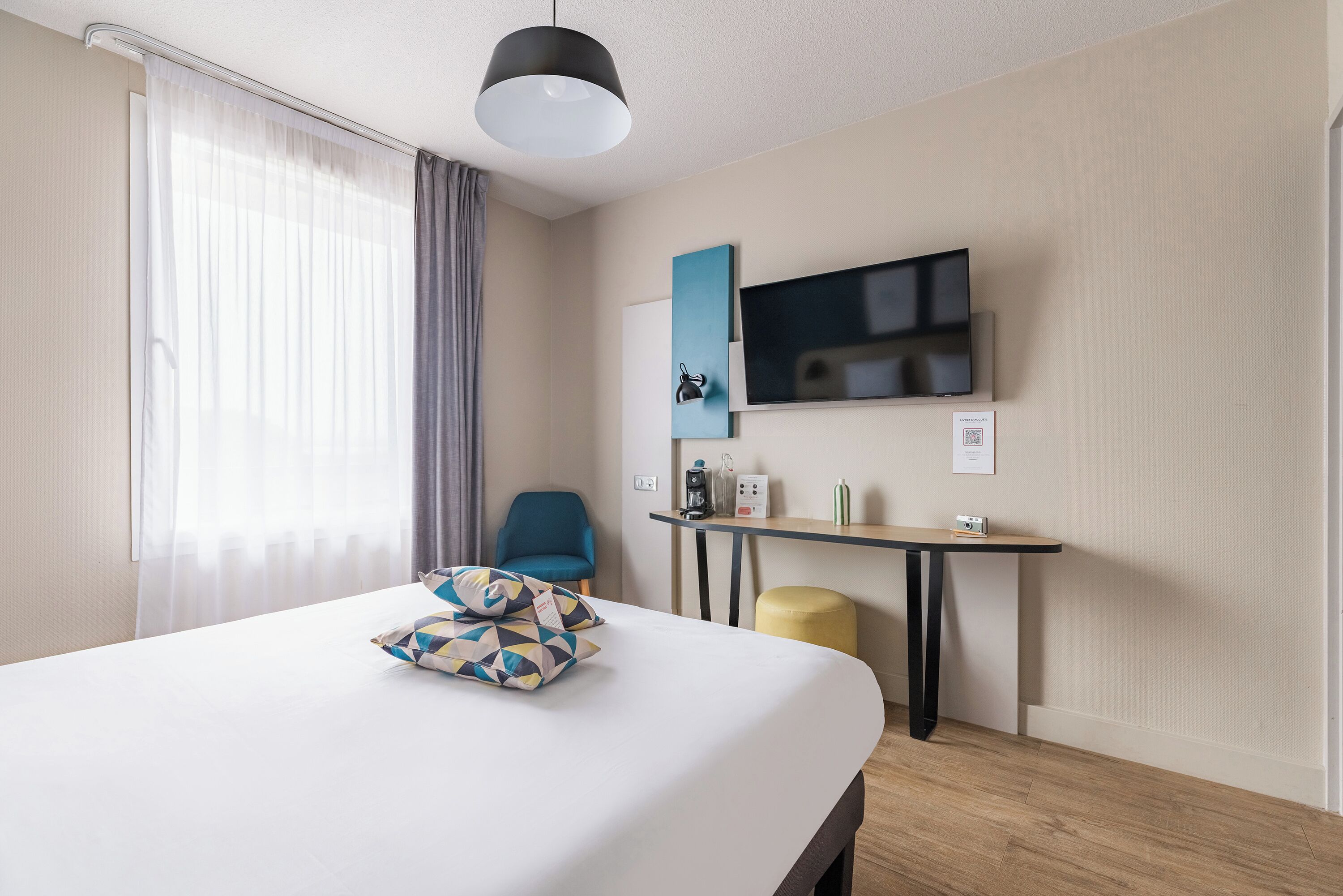 Habitación de hotel con cama, escritorio, silla, televisión y ventana con cortinas.