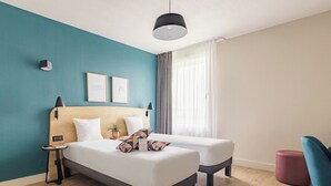 A modern hotel room with a teal accent wall, a wooden headboard, a bed with white bedding, two bedside tables, a small plant, and a blue chair. - Appart'City Confort Toulouse Diagora Labège (Labege)