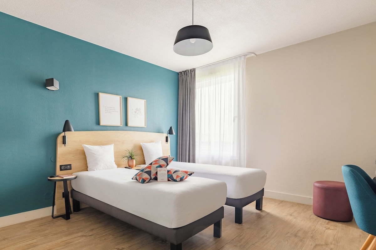 Quarto de hotel moderno com uma parede azul-turquesa, cabeceira de madeira, cama com roupa de cama branca, duas mesas de cabeceira, uma planta pequena e uma cadeira azul.