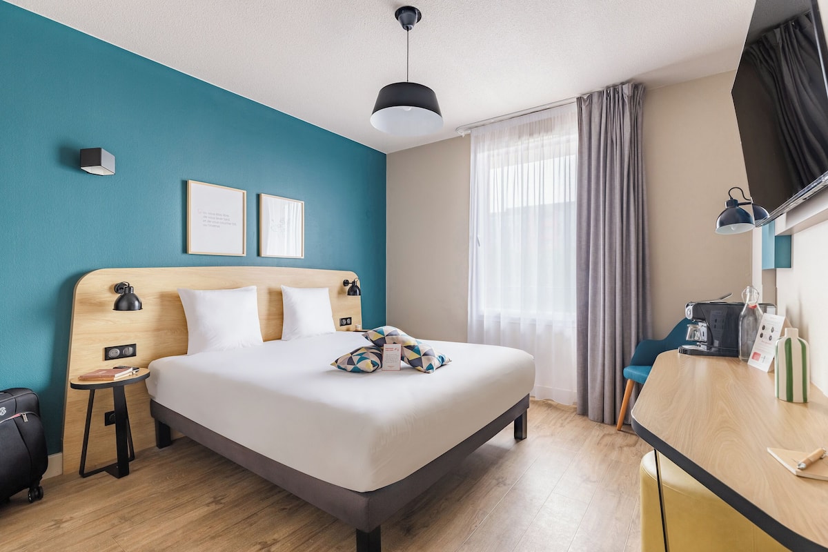 Quarto de hotel com cama, mesa de cabeceira, luminária e escrivaninha com cadeira.