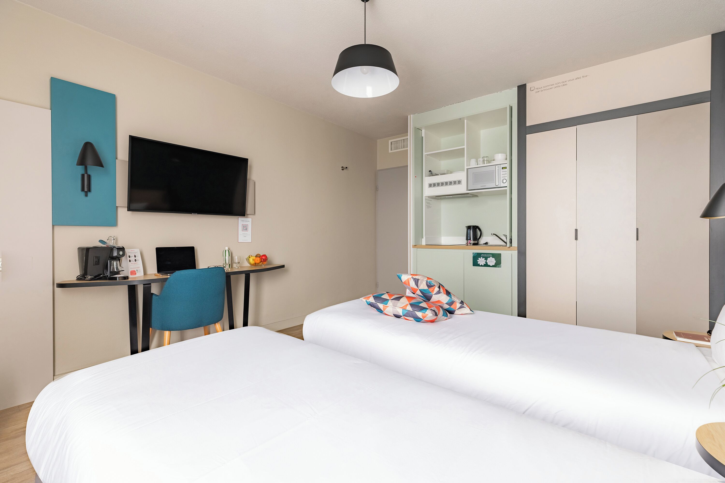 A hotel room with two beds, a desk with a computer, a TV, and a kitchenette.