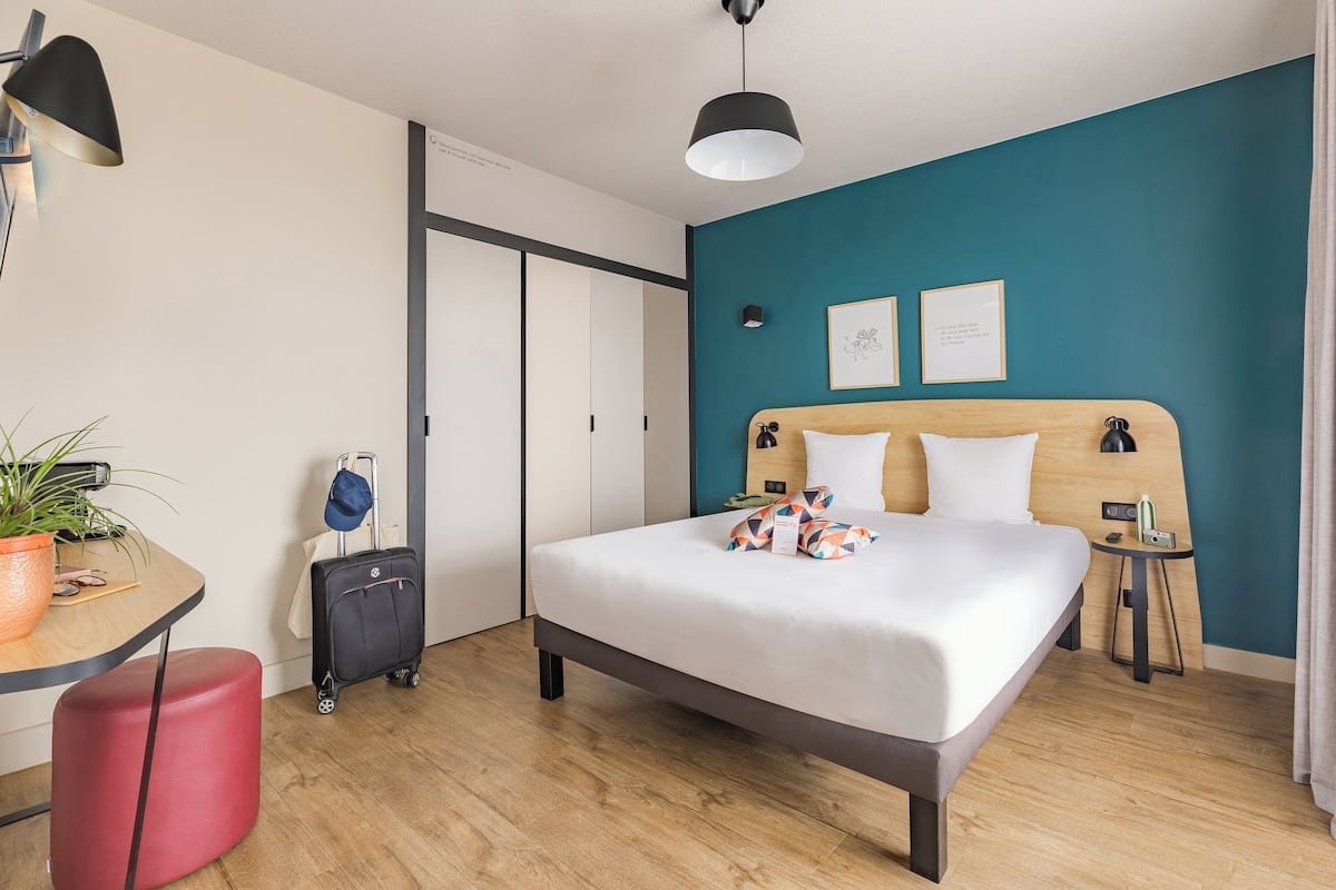 Quarto de hotel com uma cama, uma escrivaninha, um banquinho vermelho, uma mala, um abajur e uma luminária no teto.