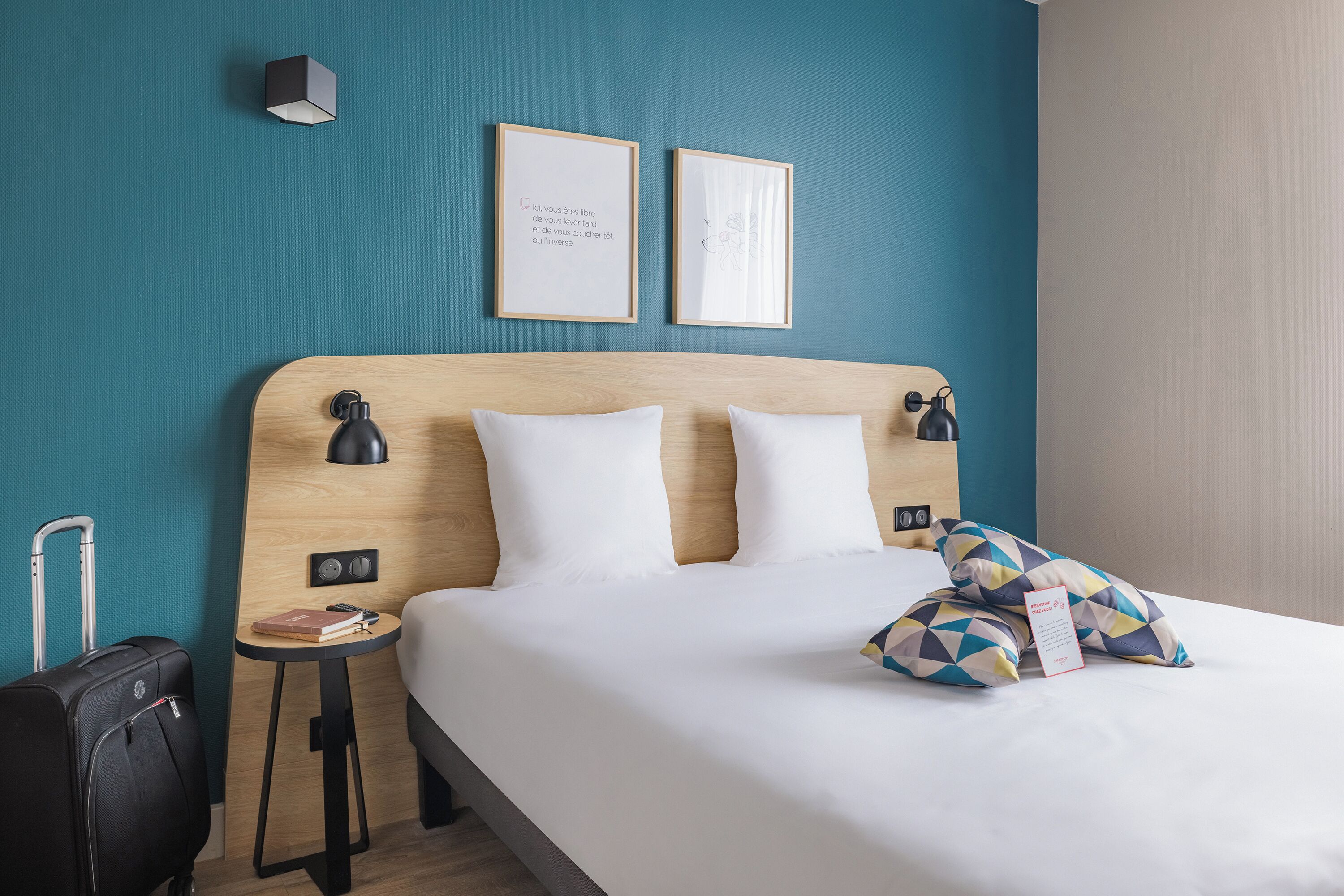 Une chambre d’hôtel avec une tête de lit en bois, du linge de lit blanc, une petite table de chevet avec un livre et une lampe, et une valise.