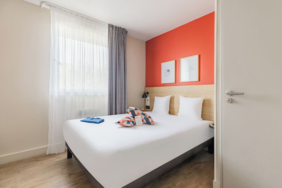 Quarto de hotel com cama, cortinas, cabeceira e duas imagens emolduradas na parede.
