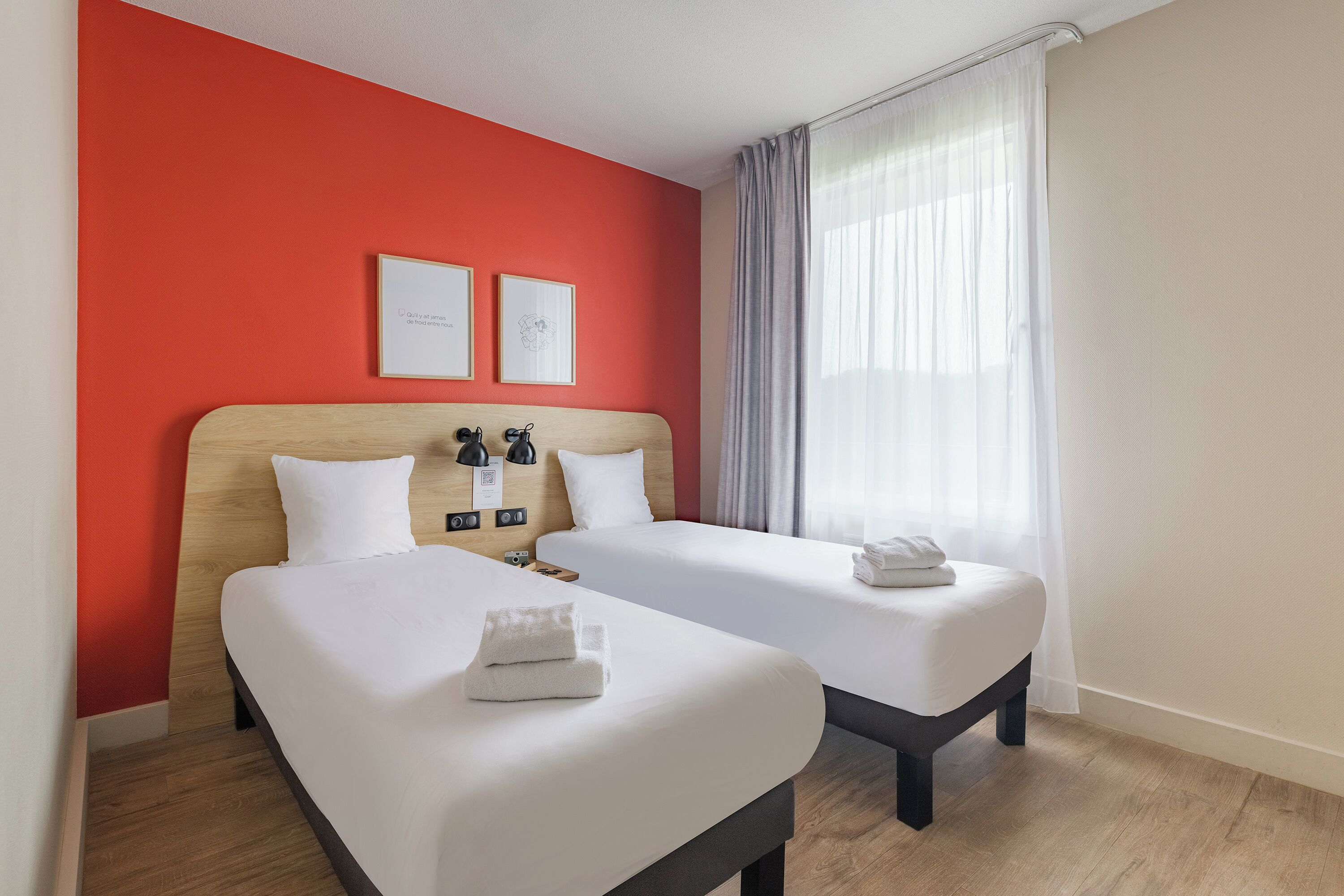 Une chambre d’hôtel avec deux lits, un mur d’accent rouge et une grande fenêtre avec des rideaux légers.