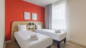 Une chambre d’hôtel avec deux lits, un mur d’accent rouge et une grande fenêtre avec des rideaux légers.
