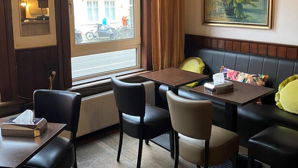 A restaurant interior with a dining area featuring black leather chairs, a wooden table, and a large window with a view of the street.
