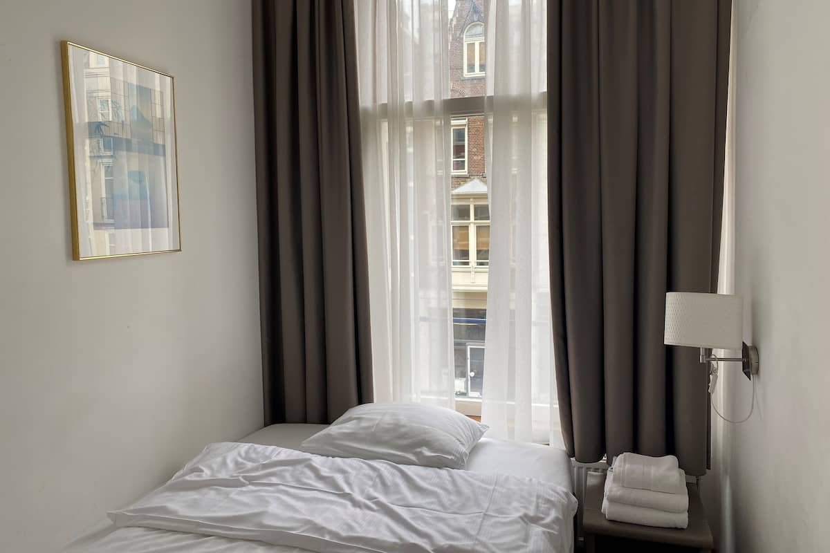 A hotel room with a bed, a framed picture on the wall, a window with curtains, and a towel rack with towels.