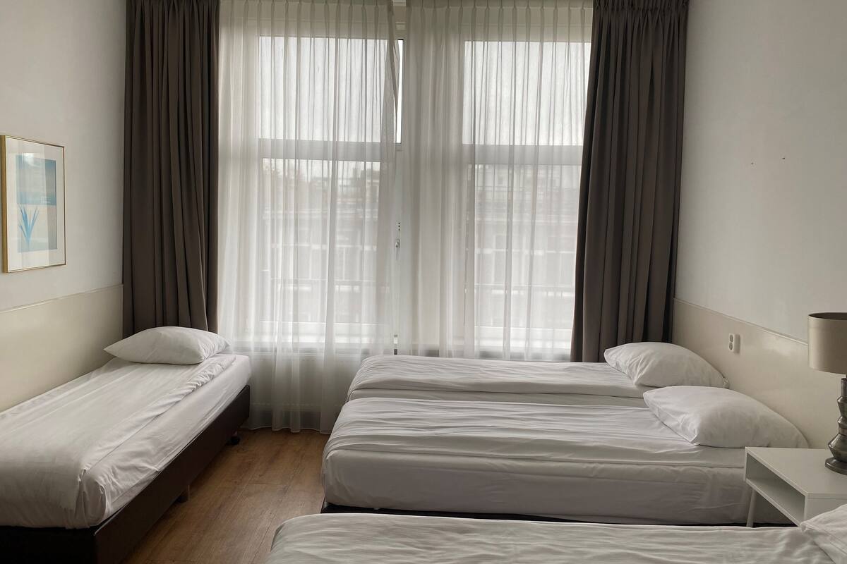 A hotel room with two single beds, a large window with sheer curtains, and a bedside table with a lamp.