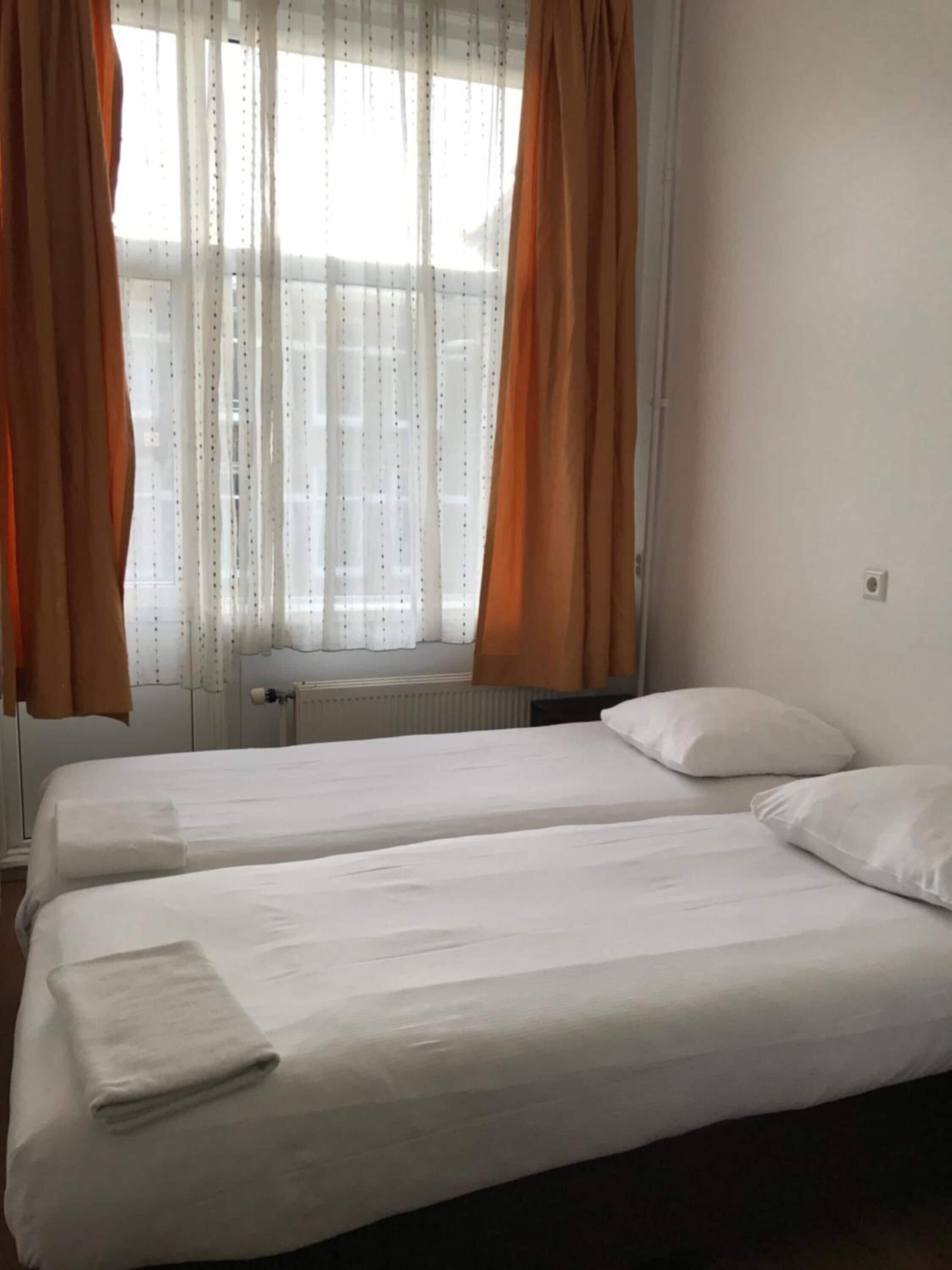 Habitación de hotel con dos camas individuales, ropa de cama blanca, un radiador y cortinas traslúcidas con cortinas color naranja.