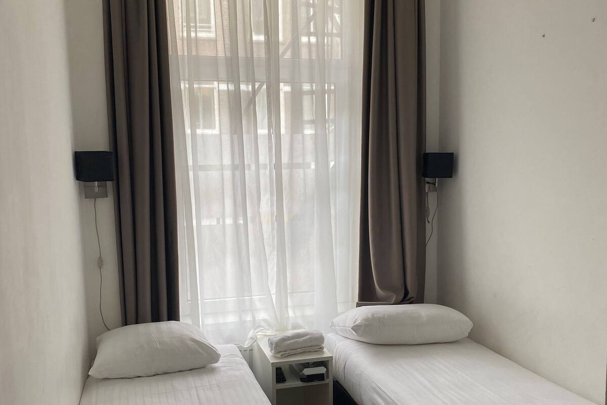 A small, clean room with two single beds, a bedside table, a nightstand, and a window with sheer curtains.