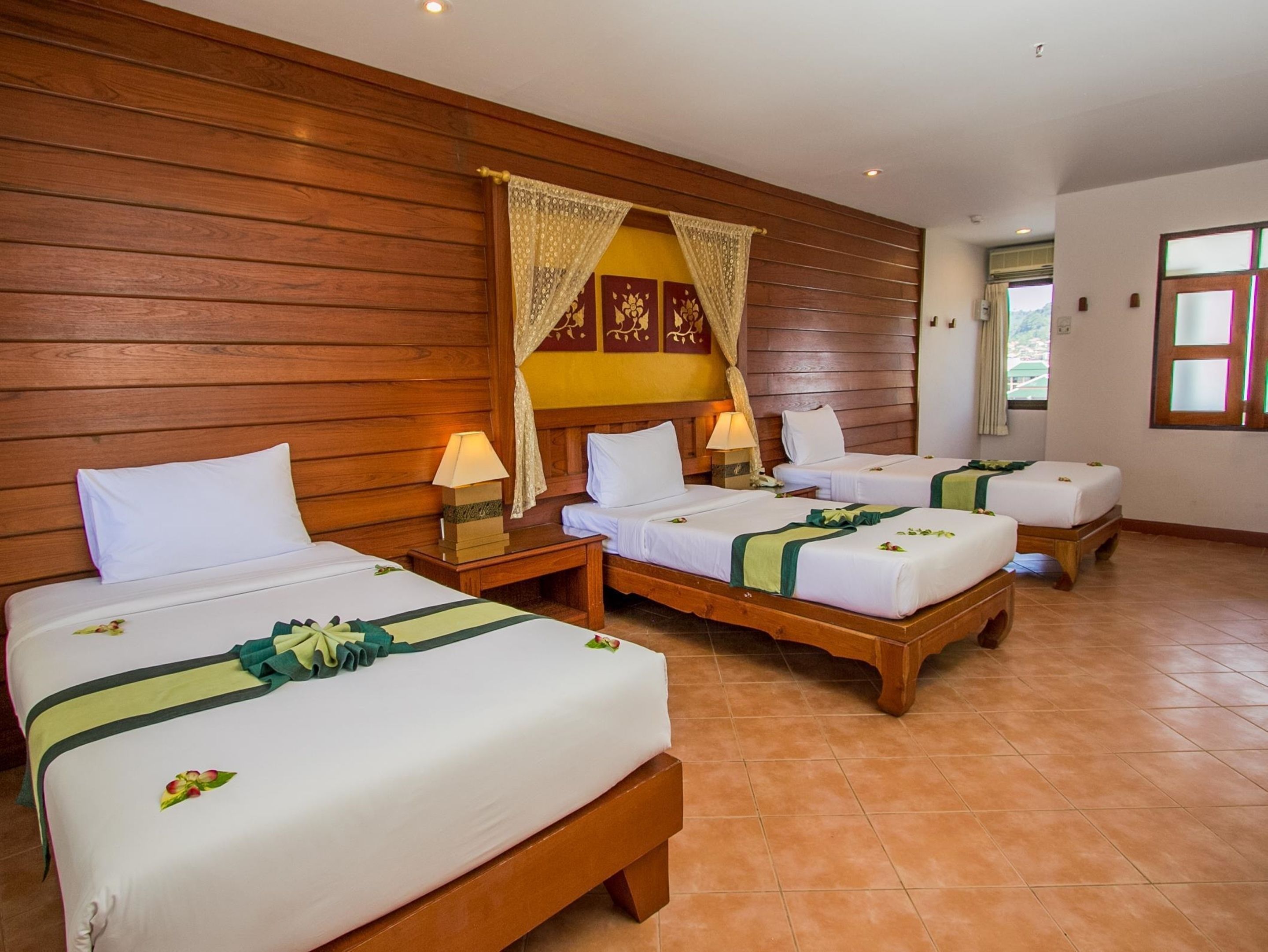 A hotel room with three wooden beds, each with white linens and green decorative pillows. There are wooden nightstands with lamps, and a wooden headboard with a decorative panel.